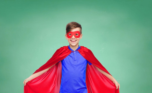 Foto concetto di scuola, educazione, infanzia, potere e persone - ragazzo felice in mantello da super eroe rosso e maschera su sfondo verde lavagna