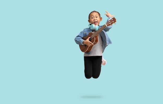 School girl Happy Asian student school kid jumping for joy with hand holding guitar ukulele Full body portrait isolated on pastel plain light blue background with clipping path for design work