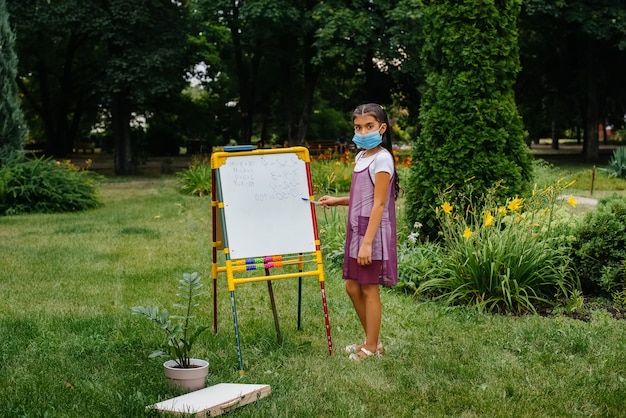 Photo a school girl in a mask stands and writes lessons on the blackboard. back to school, learning during the pandemic.