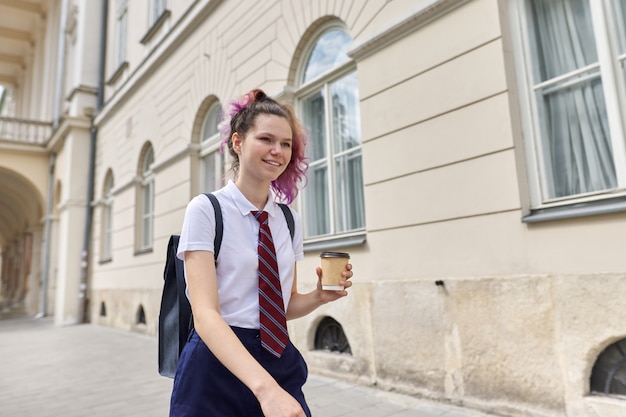 バックパックと一杯のコーヒーと歩いている学校の女の子