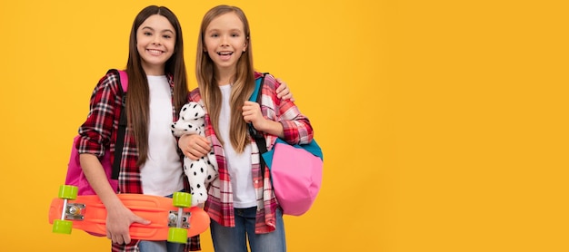 School girls friends happy kids in casual checkered shirt carry backpack toy and penny board casual