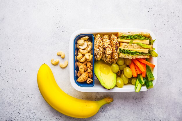 Scuola sana scatola di pranzo con sandwich, biscotti, frutta e avocado su sfondo bianco.