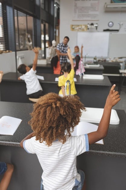バックグラウンドで教師と学校の教室で手を上げる学校の子供たち