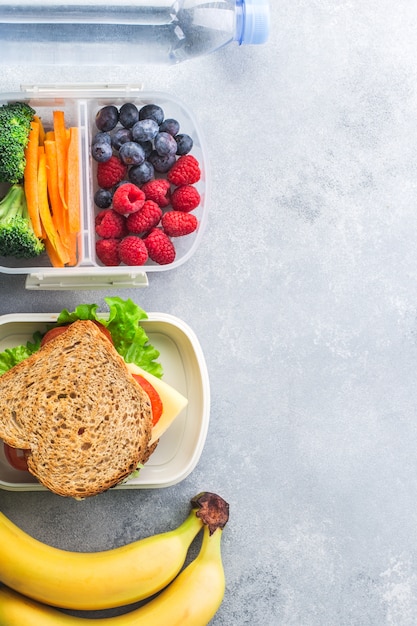 School lunch box with sandwich vegetables berries banana on grey table healthy