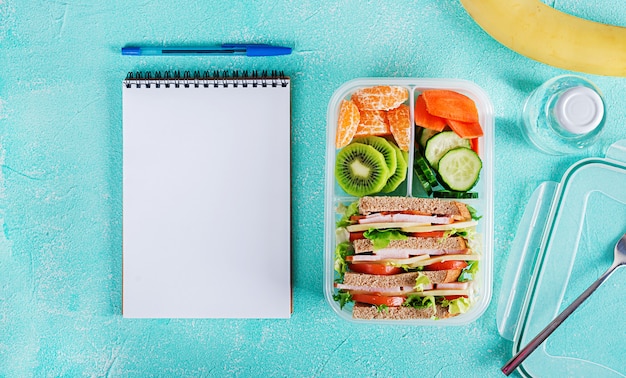 School lunch box with sandwich, vegetables, water, and fruits on table. 