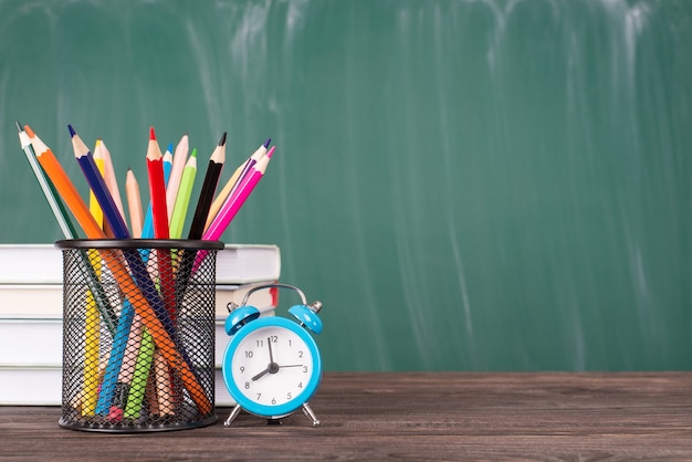 School onderwijsconcept. Close-up foto van stapel texbooks kleurpotloden en blauwe klok geïsoleerd op groen schoolbord en houten tafel copyspace