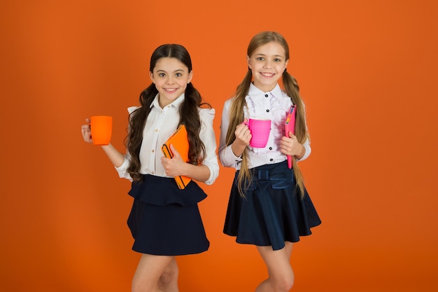 School routine Having break relax Drinking tea while break School mates relaxing with drink Enjoy being pupil Girls kids school uniform orange background Schoolgirl hold book or notepad and mug