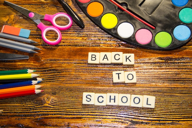 Школьный набор с надписью обратно в школу, карандашами, ручкой, ножницами, ластиком и акварельными красками на деревянном столе