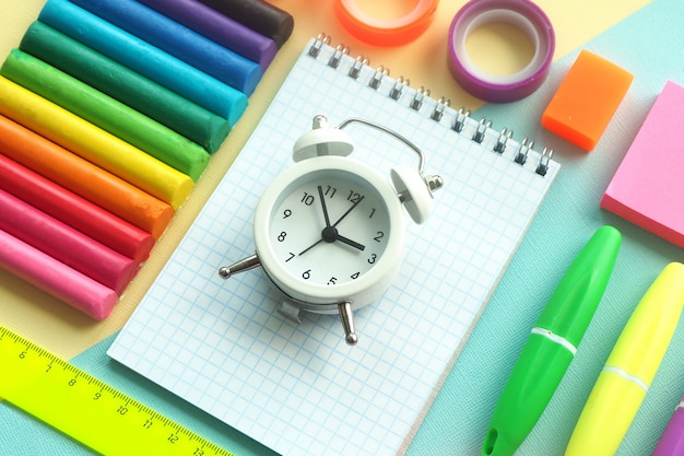 School stationery: plasticine, markers, notepad, ruler, stickers, scotch tape and a white alarm clock on a yellow-blue background.