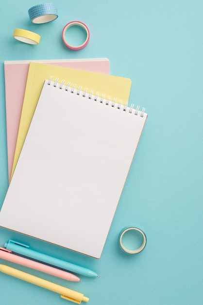 School supplies concept Top view vertical photo of colorful school accessories stack of planners pens and adhesive tape on isolated pastel blue background with empty space