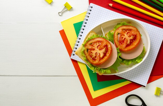 Rifornimenti di scuola e scatola di pranzo con i panini della salsiccia su un fondo di legno bianco