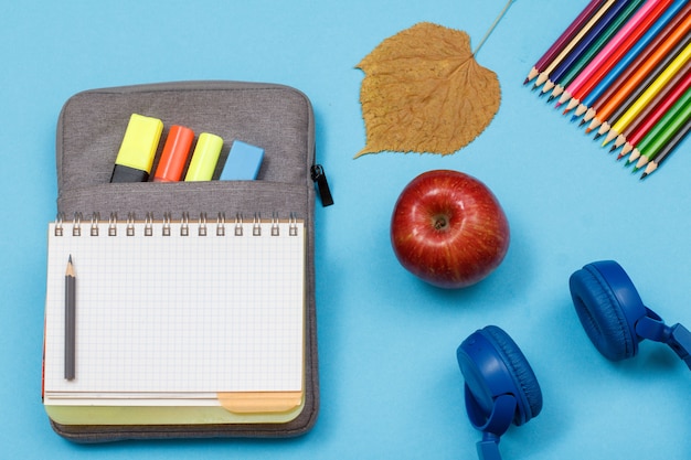 School supplies. Open exercise book on bag-pencil case with color felt pens and marker, dry leaf, color pencils, apple and headphones on blue background. Top view. Back to school concept