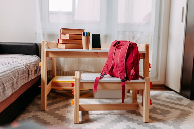 School supplies on the school desk. Red backpack, white headphones, notebook, big red books, pens in the jar lay on the white school desk.