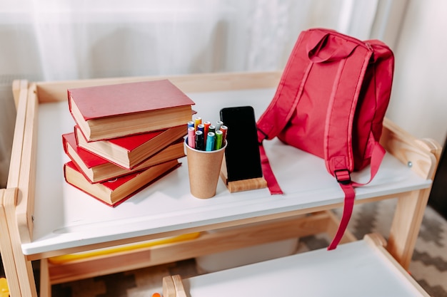 학교 책상에 학교 용품. 빨간 배낭, 흰색 헤드폰, 노트북, 큰 빨간 책, 항아리 속의 펜이 흰색 학교 책상 위에 놓여 있습니다.