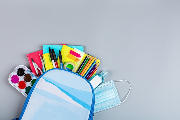 School supplies with medical face mask on a gray background. Protection of schoolchildren and students from the coronavirus covid-19, schooling in a pandemic. Flat lay, top view, mockup, template