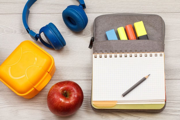 Foto materiale scolastico. scatola di sandwich gialla, mela, cuffie, quaderno aperto su astuccio borsa-matita con pennarelli colorati e matita su tavole di legno grigie. vista dall'alto con copia spazio. ritorno al concetto di scuola