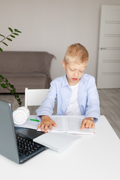 Schoolboy boy blonde learns to read a book during distance learning at home via laptop, concept back to school