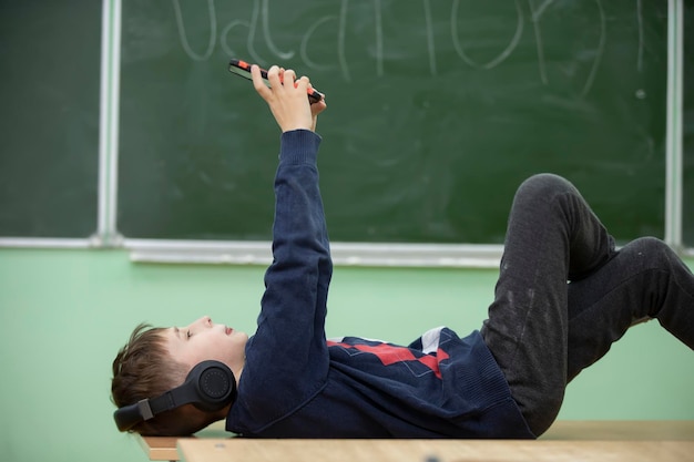 A schoolboy boy lies in headphones on a school desk and looks into a smartphoneChange at school