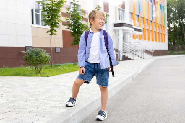 A schoolboy boy stands with a backpack near the school smiles laughs elementary school