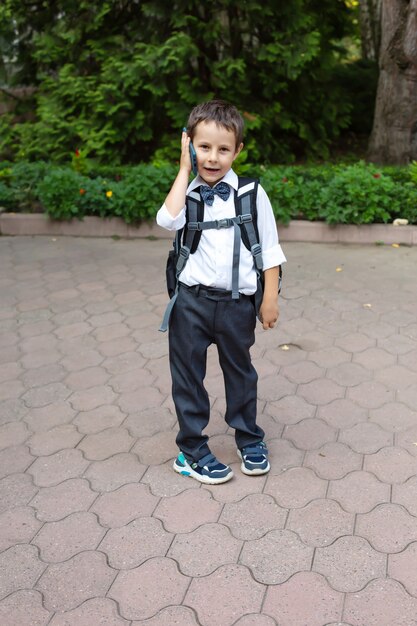 a schoolboy boy in a white shirt gray trousers with a backpack holds a phone