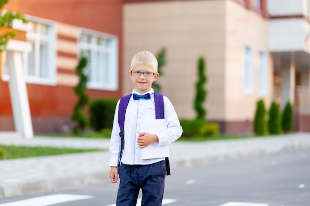 A schoolboy boy with blond glasses with a backpack and a white book is standing at the school