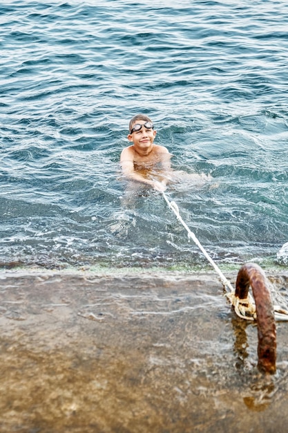 Schoolboy catches and holds rope swimming in blue water trying to climb up stone pier