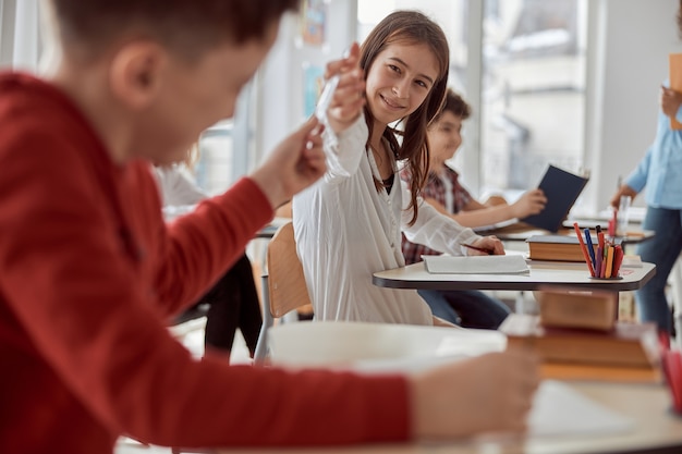 Schoolboy gives pencil to his classmate while sitting at desk while teacher speaking in school classroom.