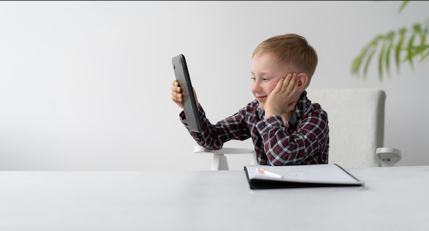 A schoolboy is doing quarantine homework at home. Distance learning child. The boy is sitting in front of the table. On the table is a large notebook.. Skype lessons. Online education at