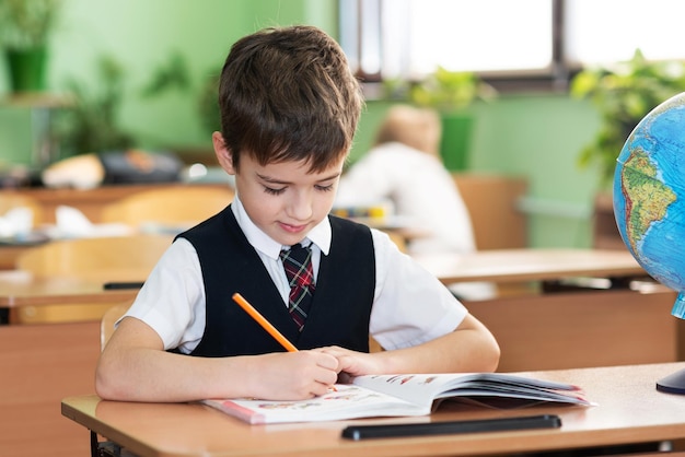 A schoolboy is sitting at a desk in an empty classroom and is writing something in a notebook