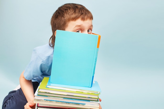 Schoolboy is sitting with a stack of books and reading and covers his face with a book