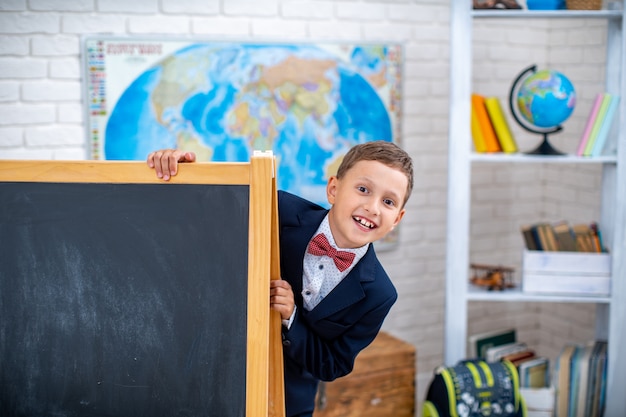 Schoolboy looks out from behind black blackboard in the classroom.