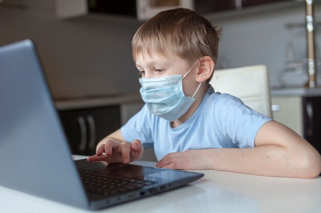 Schoolboy in a medical mask dresses at home, does homework at home. The boy looks at the laptop. Back to school. Distance Learning Online Education