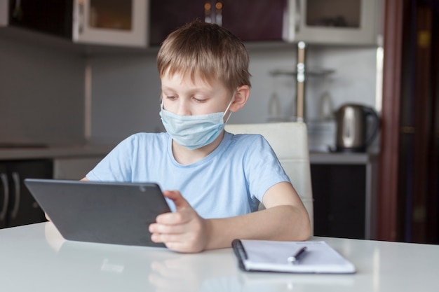 Lo scolaro in una mascherina medica si veste a casa, fa i compiti a casa. il ragazzo guarda il tablet. di nuovo a scuola. formazione online a distanza