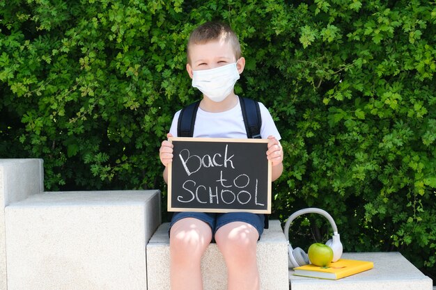 Schoolboy in a protective mask from the virus with a sign in his hands stands on the street on the way to school. Back to school