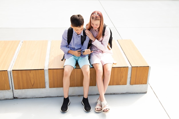 Photo schoolboy schoolgirl teenagers laugh have fun after lessons sitting on wooden bench in schoolyard, concrete background use tablet, concept of online education, modern technologies in kids life