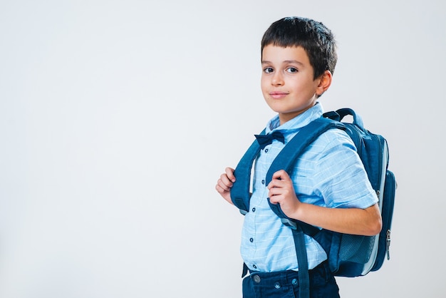 The schoolboy in a shirt with a bow tie and a backpack stands half a turn behind his back and smiles