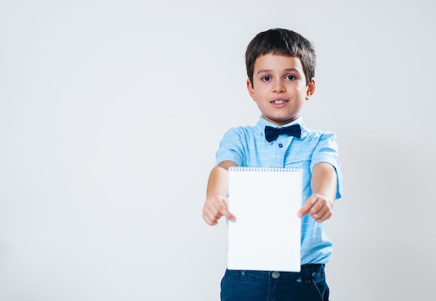 The schoolboy in a shirt with a bow tie holds a notebook in front of him and points it to the camera