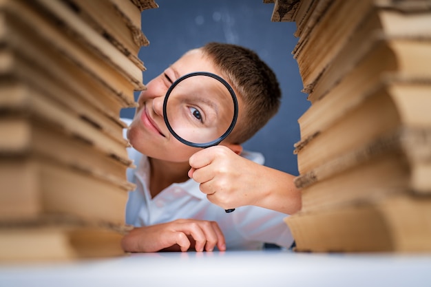 Photo schoolboy sitting between two piles of books looking at camera through the magnifying glass.