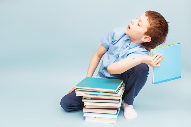 Schoolboy sitting with pile of school books and throwing a textbook isolated on a blue background