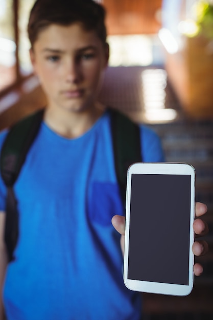 Schoolboy standing with schoolbag showing mobile phone near staircase at school