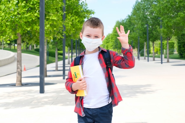 Schoolboy wearing a protective mask against the virus, with a textbook in his hands, stands on the street on his way to school. Back to school