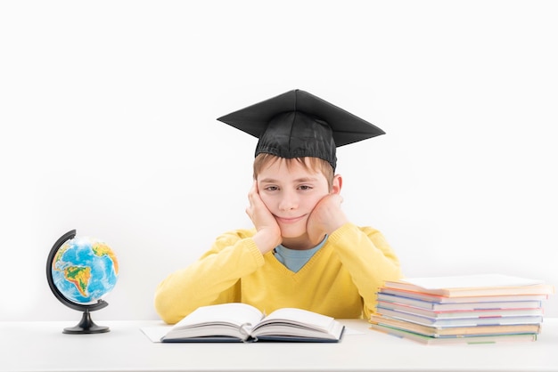 Schoolboy wears the students hat and sitting at desk in class Portrait of student on white background