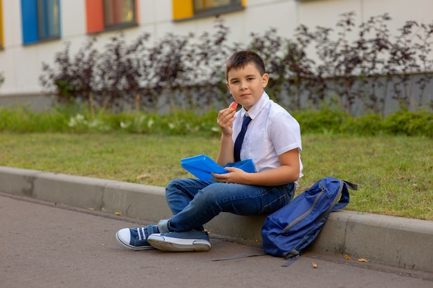 Школьник в белой рубашке с синим галстуком, держит синюю ланч-бокс и дольку яблока, смотрит в камеру