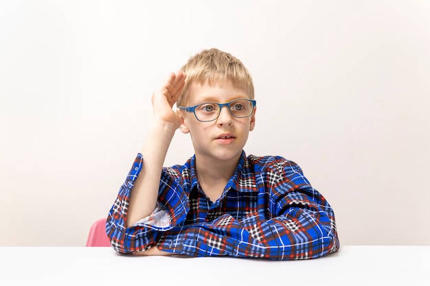 Schoolboy with glasses raises his hand on a white table and white wall background