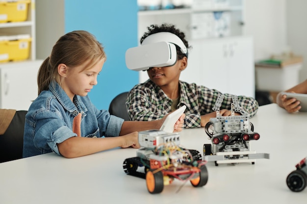 Schoolboy with vr headset turning head towards his classmate with tablet