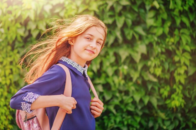 A schoolgirl in a blue dress with a white collar and a backpack on a background of green foliage