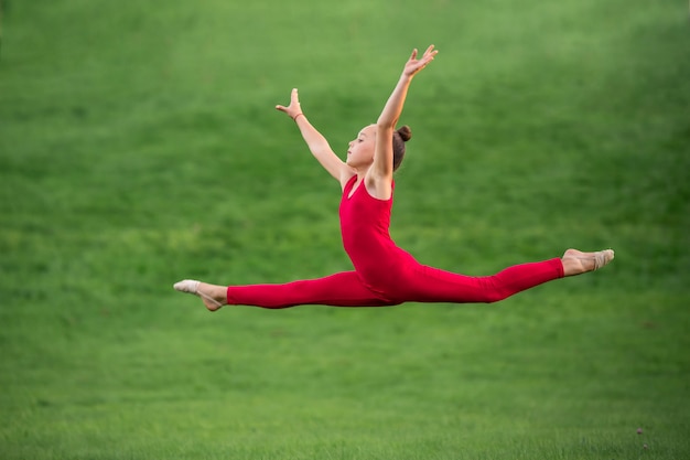 Schoolgirl in bright red overalls is engaged in gymnastics on the grass, doing exercises for stretching