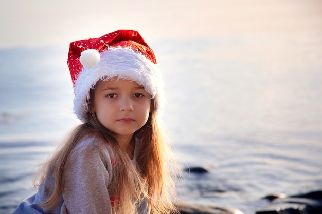 A schoolgirl girl in a New Year's hat sits on the seashore. Christmas in warm countries. Vacation and New Year's holidays at the sea