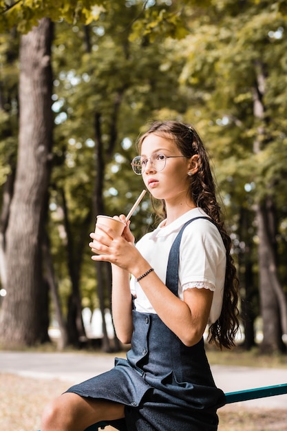 A schoolgirl in glasses drinks a beverage from an eco paper cup with a straw in the park