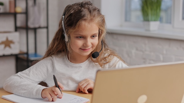 Schoolgirl in headset has online lesson, video call with teacher. Cute primary school girl studying at home using laptop.